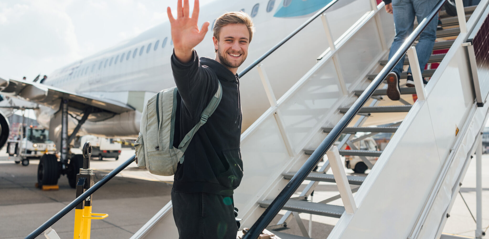 A young backpacker getting onto a plane while budget travelling. He is walking up the steps to the plane carrying his suitcase, smiling at the camera while waving.